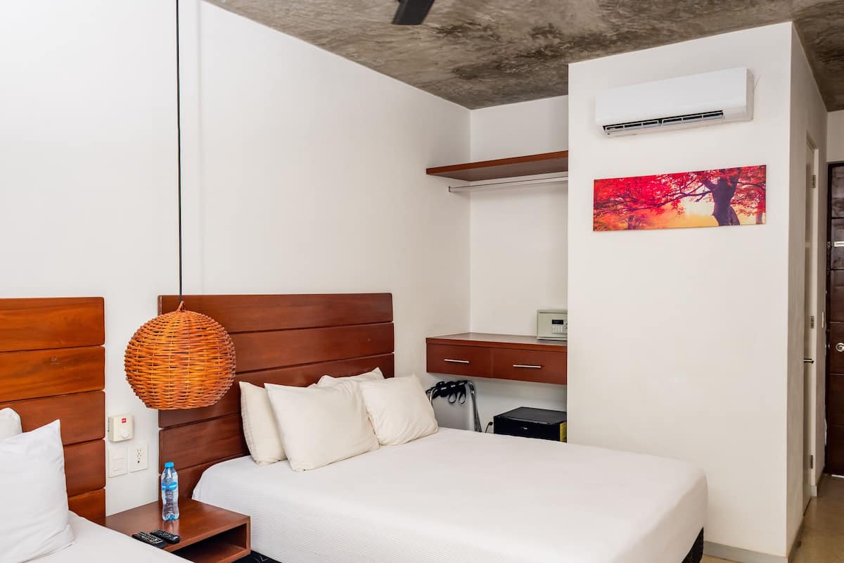 A hotel room with a wooden headboard, a bed with white bedding, a bedside table with a bottle and remote control, a wall-mounted air conditioner, a painting, and a wooden door.