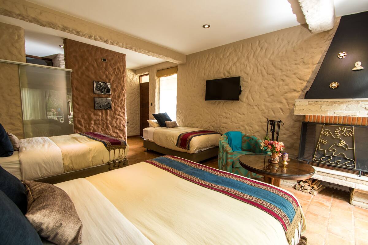 Quarto de hotel com duas camas, um sofá, uma lareira e uma televisão na parede.