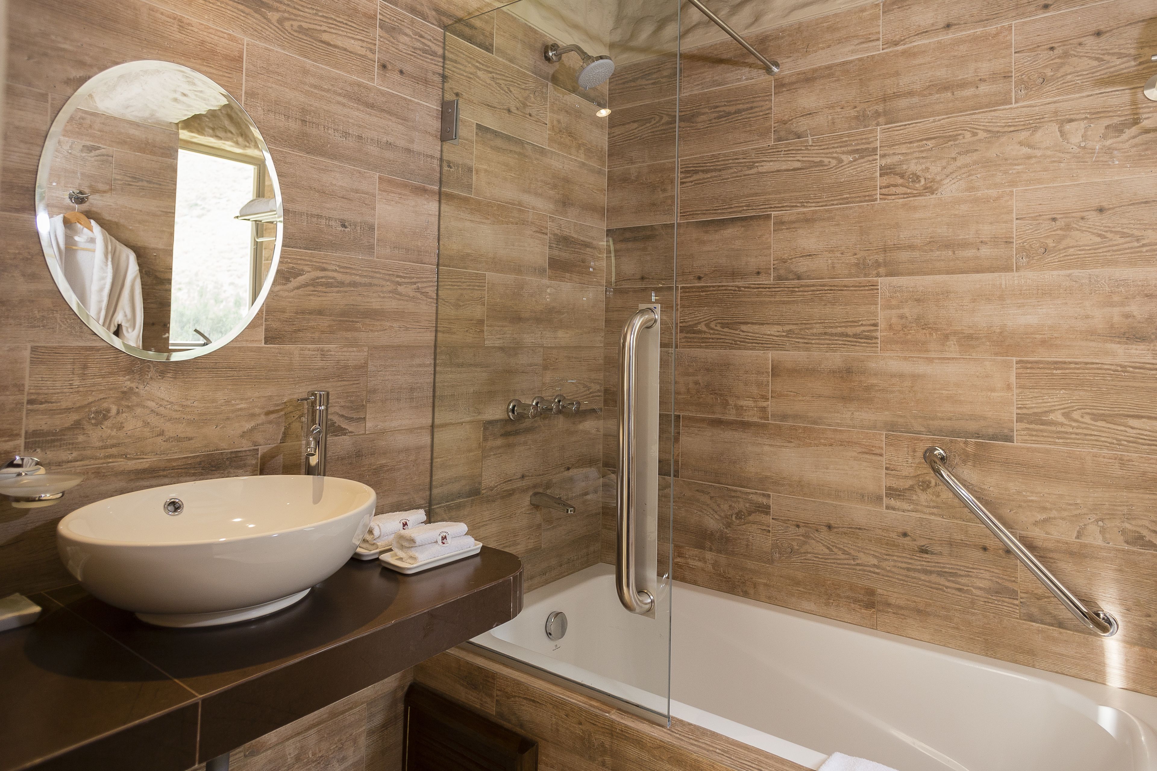 Ein modernes Badezimmer mit Holzboden, rundem Spiegel, weißem Waschbecken und einer barrierefreien Dusche mit Haltegriffen.