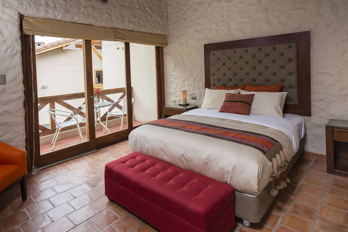 Quarto com cama grande, um banco vermelho, porta de vidro deslizante para área externa e cabeceira de madeira.