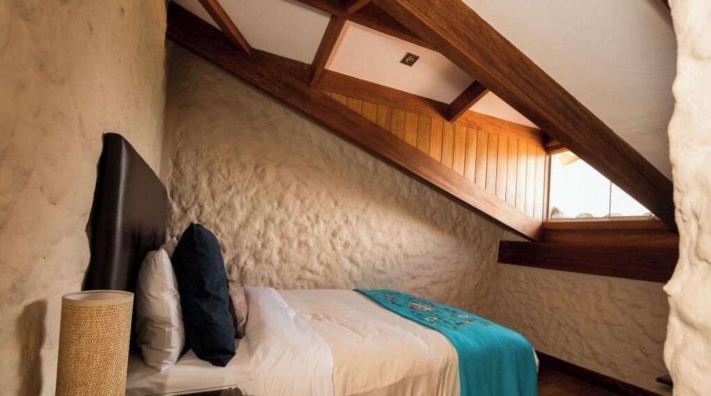 Une chambre avec un plafond en pente, un lit recouvert d’une couverture bleue et un panier en osier.