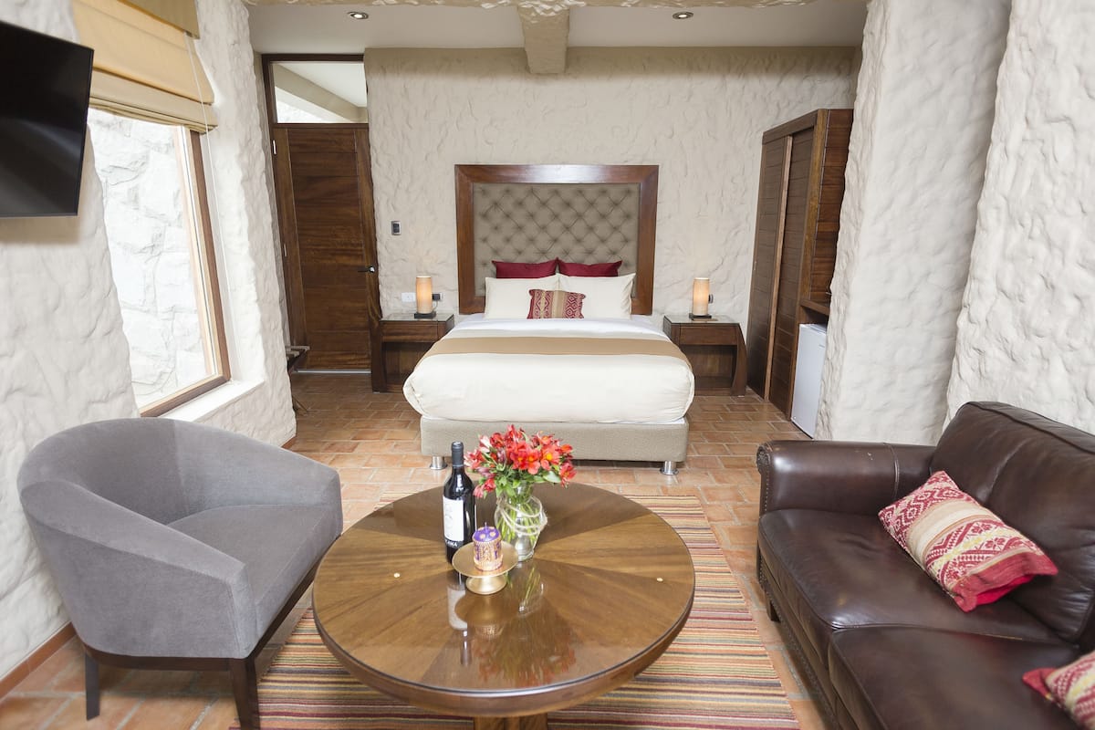 Quarto de hotel com uma cama, uma poltrona cinzenta, um sofá de couro marrom, uma mesa de centro redonda com um vaso de flores e uma garrafa de vinho.
