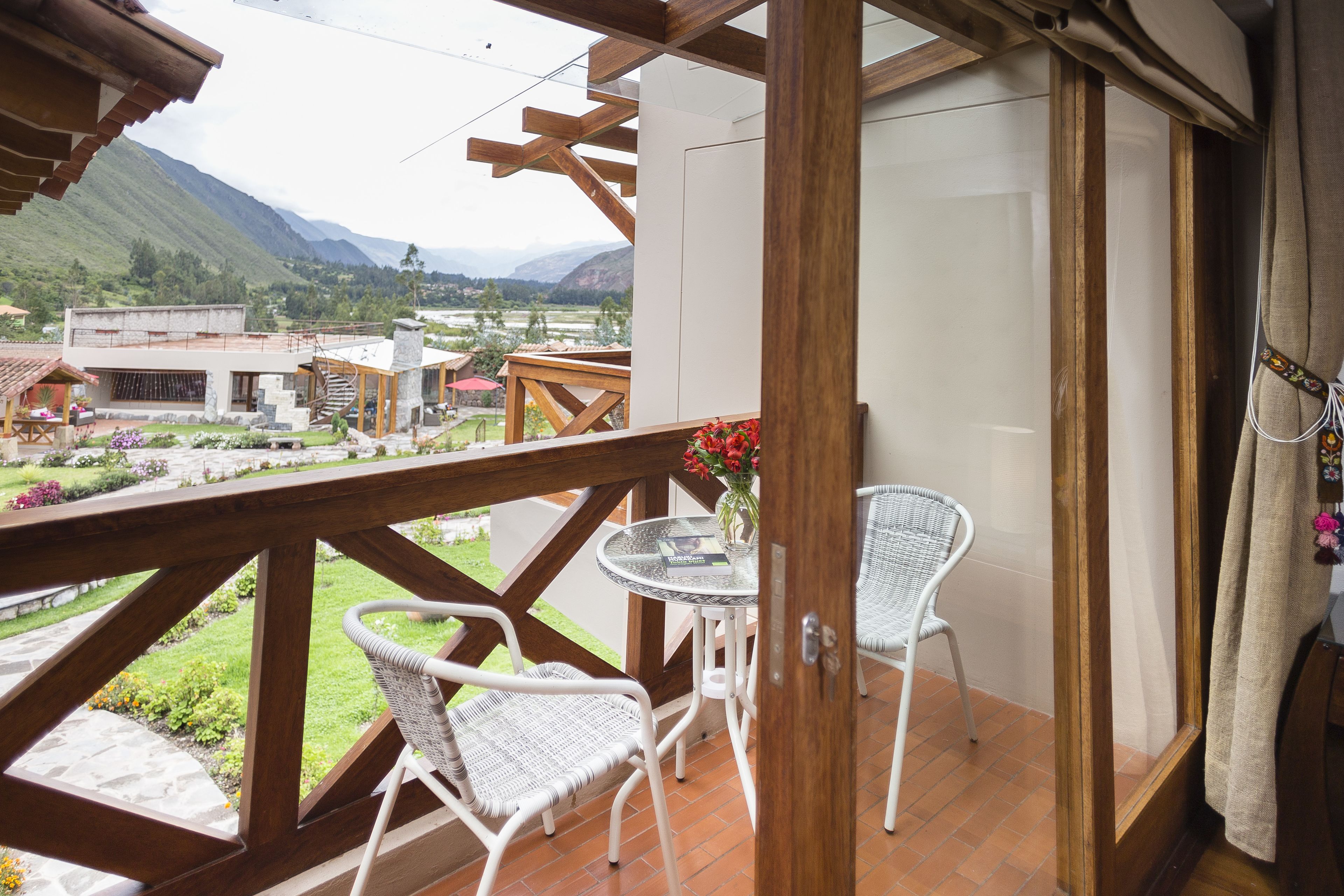Un balcón con sillas de mimbre blanco, una pequeña mesa redonda con un jarrón de flores y vistas a un paisaje montañoso.