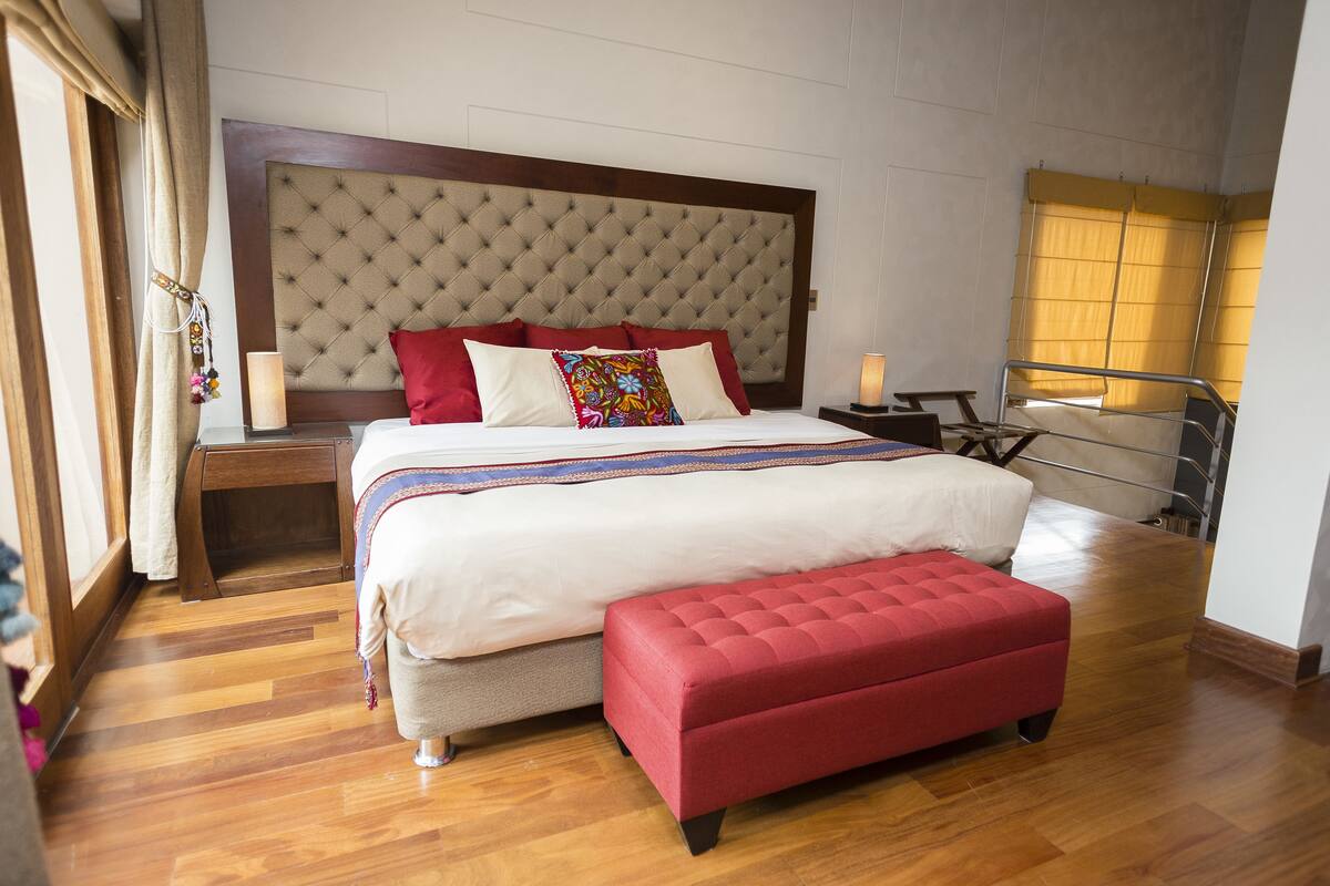 Quarto de hotel com cama grande, um banco vermelho, piso de madeira, cabeceira e uma janela com cortinas.