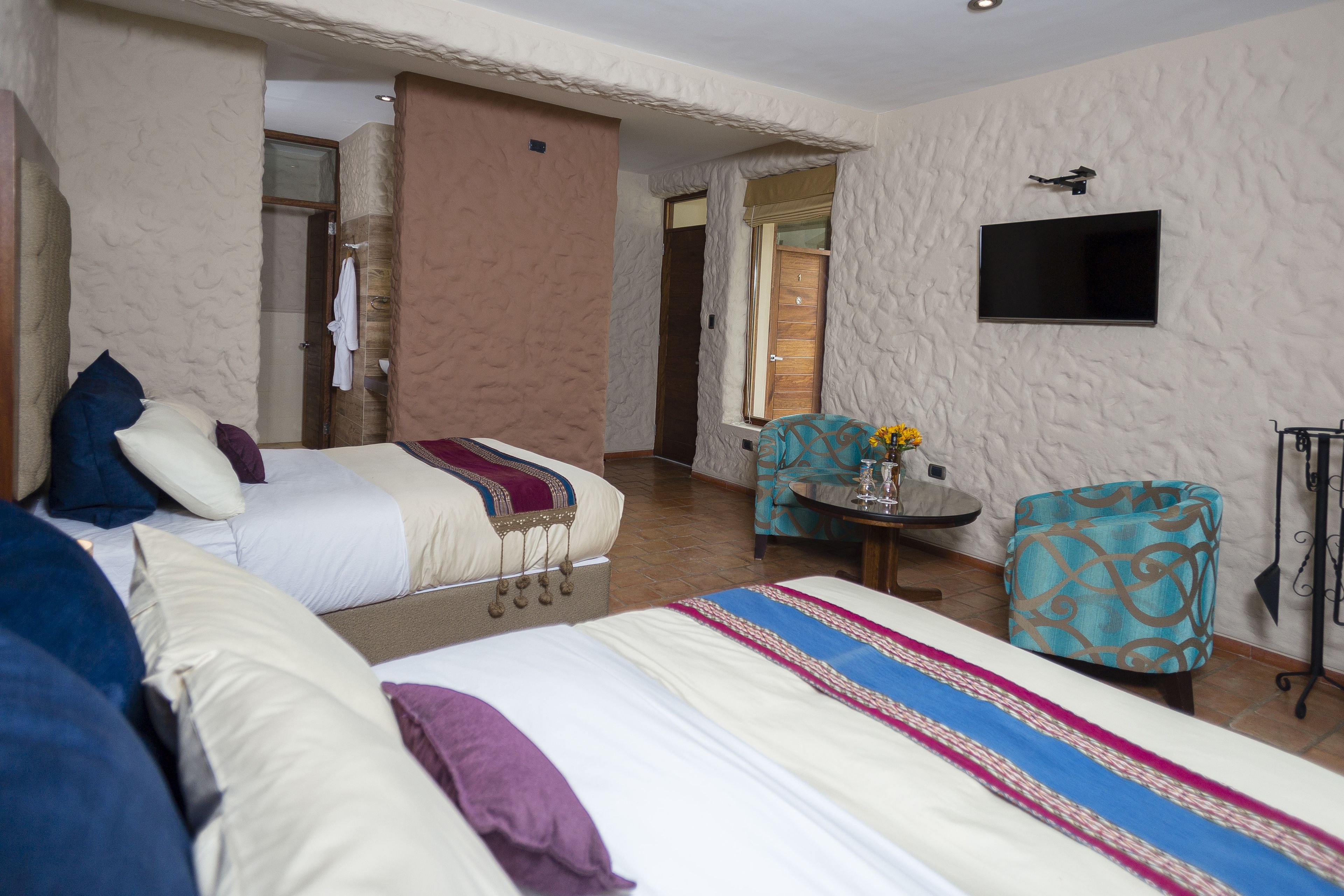 A hotel room with two beds, a TV mounted on the wall, a small table with a vase of flowers, and a patterned armchair.