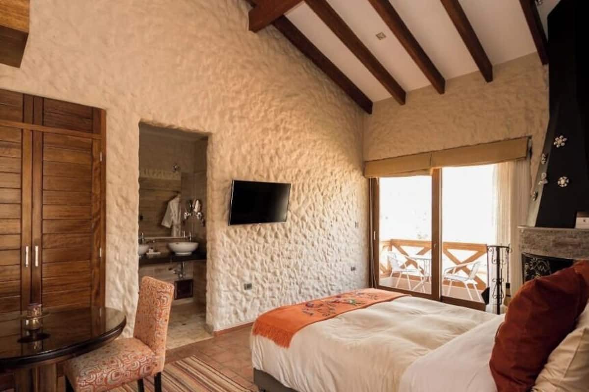 Quarto com parede de pedra, cama com travesseiro vermelho, guarda-roupa de madeira, televisão e varanda com vista.