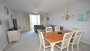 A dining area with a wooden table and white chairs, a television on a stand, a ceiling fan, and a chandelier. - Ironwood Apartment 1722 (North Myrtle Beach)