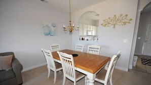 A dining area with a wooden table and white chairs, a chandelier, and a kitchen in the background. - Ironwood Apartment 1722 (North Myrtle Beach)