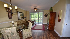A living room with a glass coffee table, patterned armchairs, a wooden cabinet, and a ceiling fan. - River Crossing Apartment 312 (North Myrtle Beach)