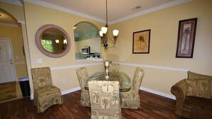 A dining area with a glass table surrounded by chairs with palm tree patterns, a round mirror, and a chandelier. - River Crossing Apartment 312 (North Myrtle Beach)