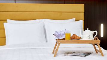 A neatly made bed with white linens and a wooden headboard. A wooden tray with a teapot, a croissant, and a glass of orange juice is placed on the bed.
