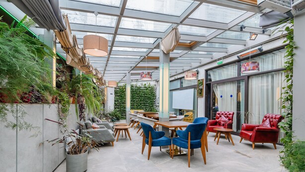 Un espace intérieur moderne doté d’un plafond en verre, de tables en bois, de chaises bleues et rouges, et de plantes en pot.