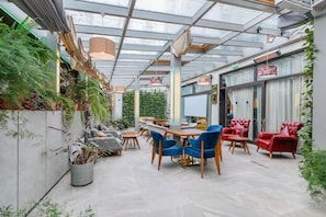 Un espace intérieur moderne doté d’un plafond en verre, de tables en bois, de chaises bleues et rouges, et de plantes en pot.