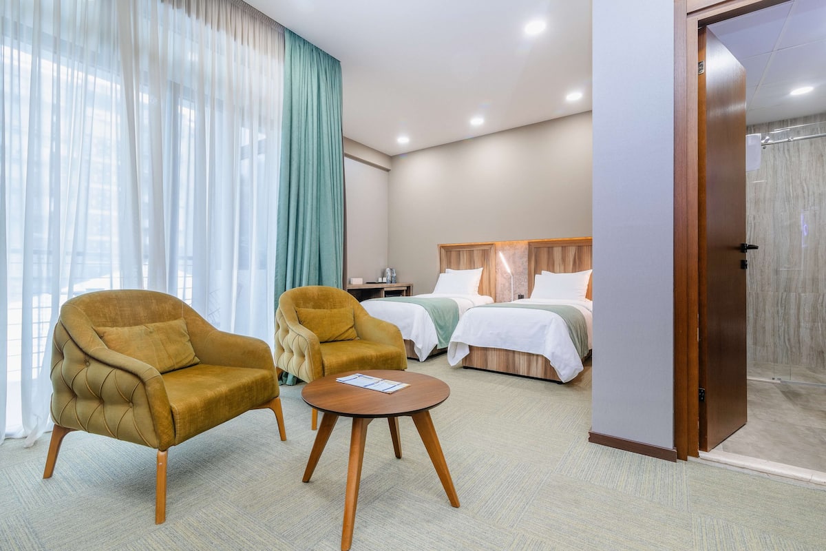 Ein Hotelzimmer mit zwei Sesseln, einem runden Holztisch, einem Bett mit Holz Kopfteil und einer Duschkabine aus Glas.