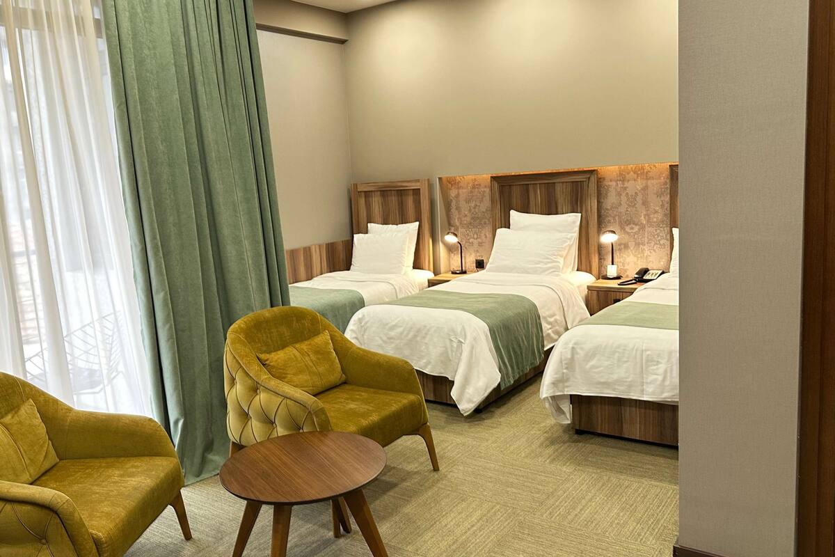 Ein Hotelzimmer mit zwei Betten, einem grünen Sessel, einem kleinen runden Tisch und einem Fenster mit durchsichtigen Vorhängen.