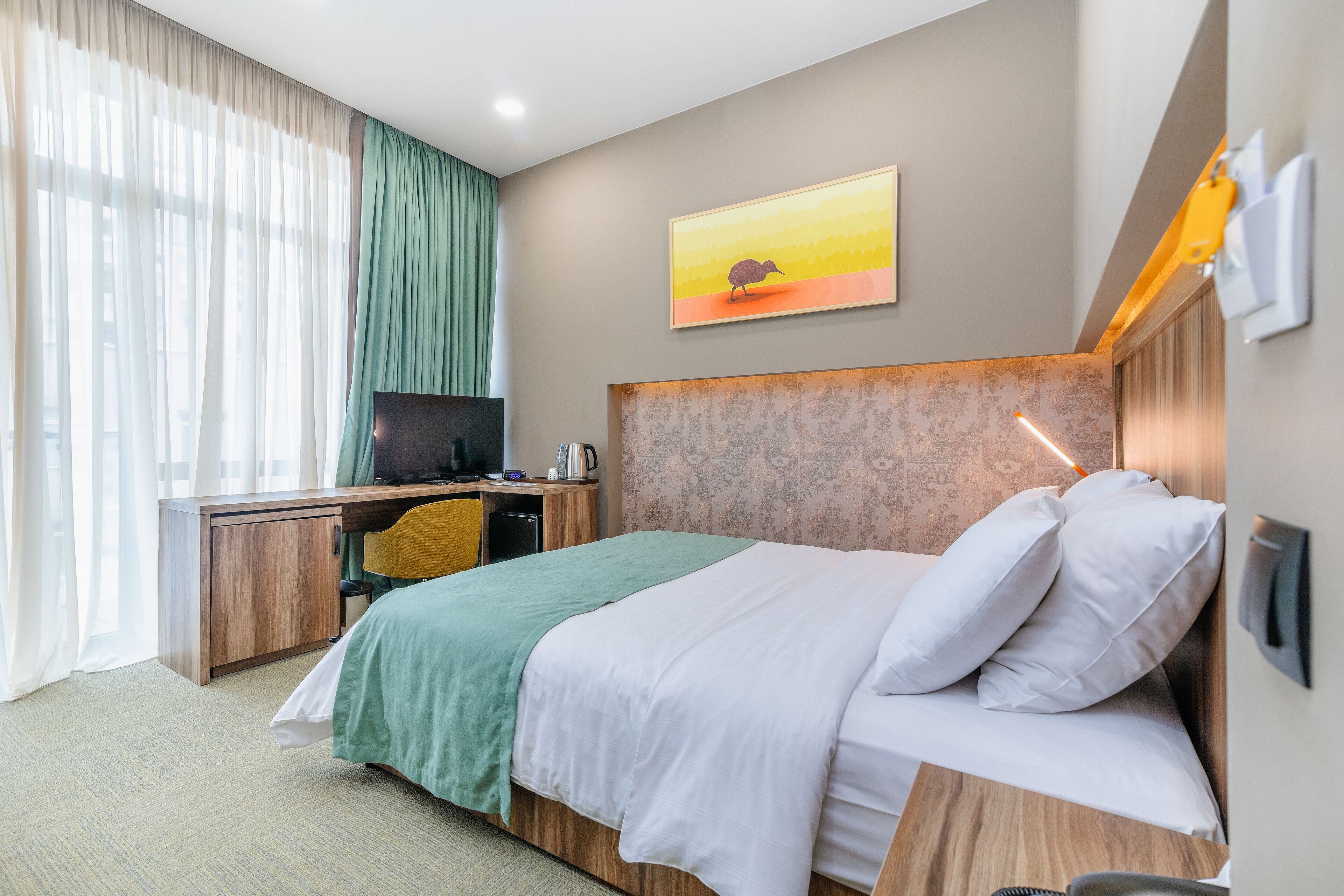 A hotel room with a bed, desk, and chair, a painting on the wall, and a window with curtains.