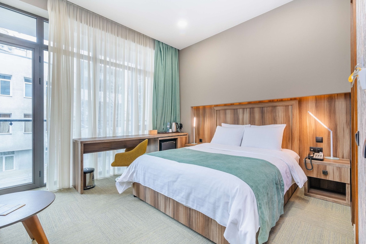 Una habitación de hotel con cama, mesita de noche, escritorio y ventana con cortinas.