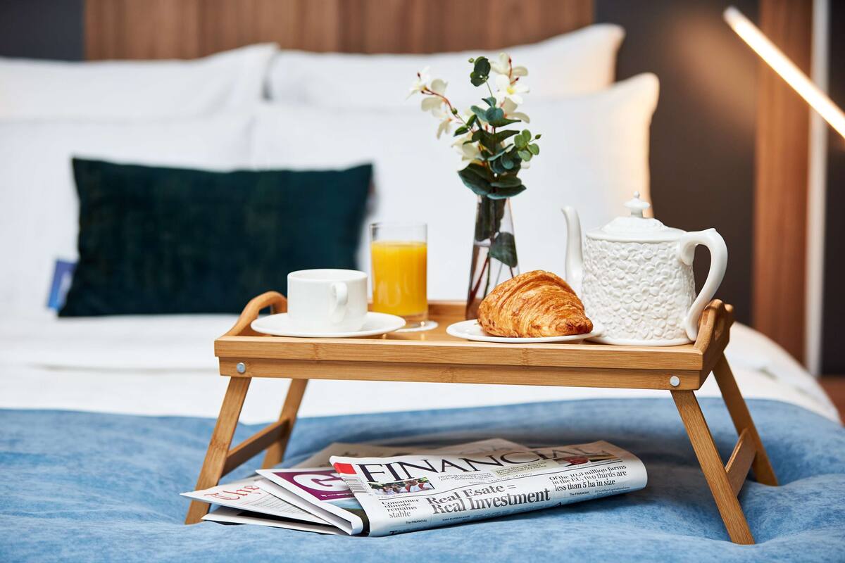 Ein Frühstücksbrettchen mit Croissant, Kaffee und Orangensaft auf einem Bett mit Zeitschrift, Blumen in einer Vase und einer Nachttischlampe.