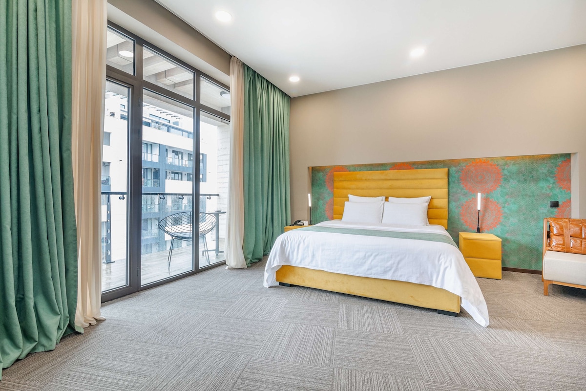 Habitación de hotel con una cama grande, una mesita de noche, una silla y vistas al exterior a través de un ventanal.