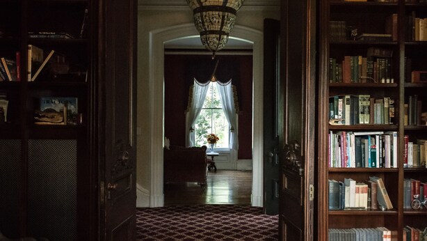 A hallway with a patterned carpet, a chandelier, and bookshelves on either side. - The Bevin House Bed & Breakfast (East Hampton)