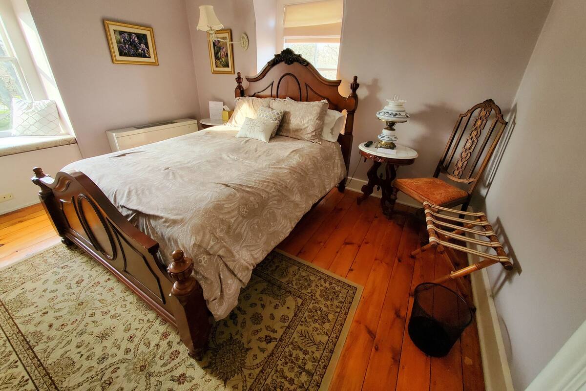 A bedroom with a wooden bed, a small round table with a lamp, a chair, and a window with a view.