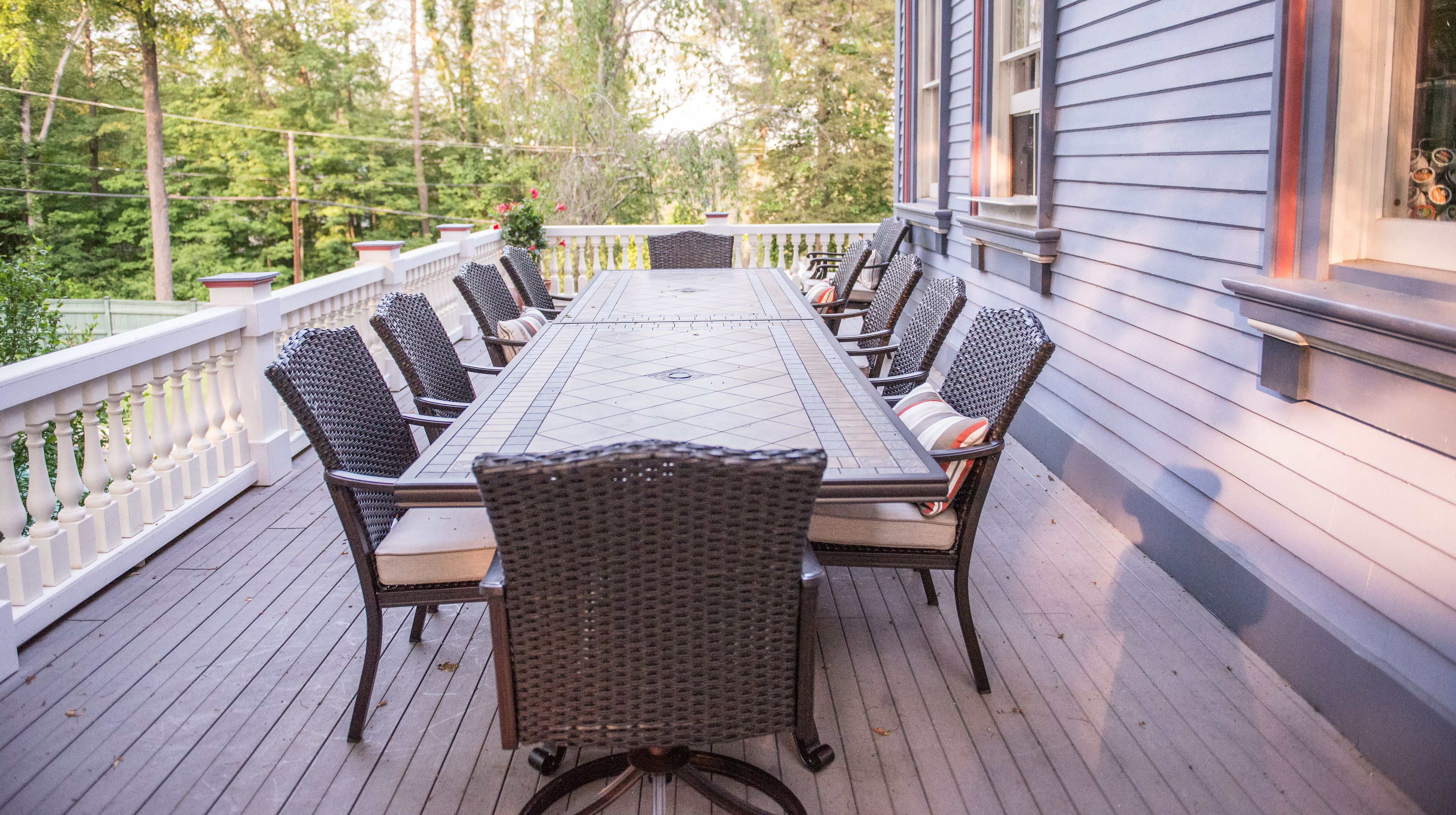 A covered outdoor dining area with a long table and wicker chairs, surrounded by a white railing and a wooden deck.