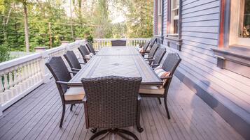 A covered outdoor dining area with a long table and wicker chairs, surrounded by a white railing and a wooden deck.