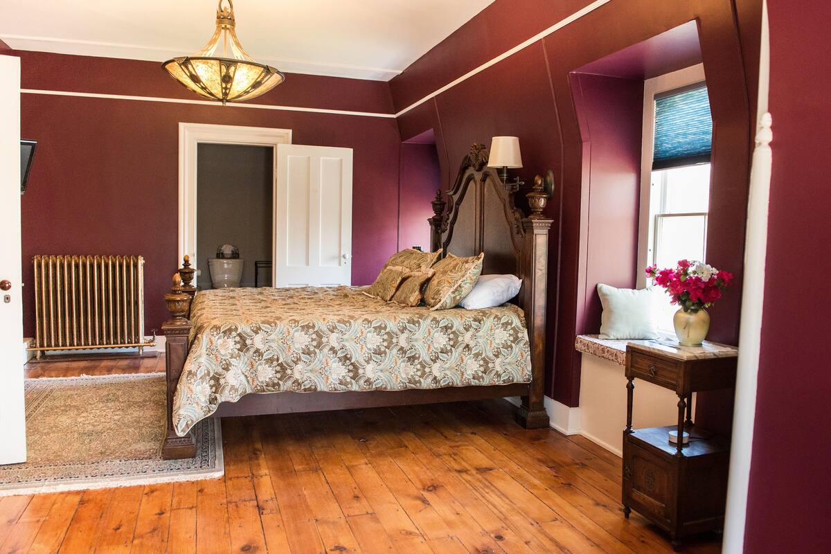 A bedroom with a wooden floor, a bed with a floral bedspread, a nightstand with a vase of flowers, a radiator, and a window with blinds.
