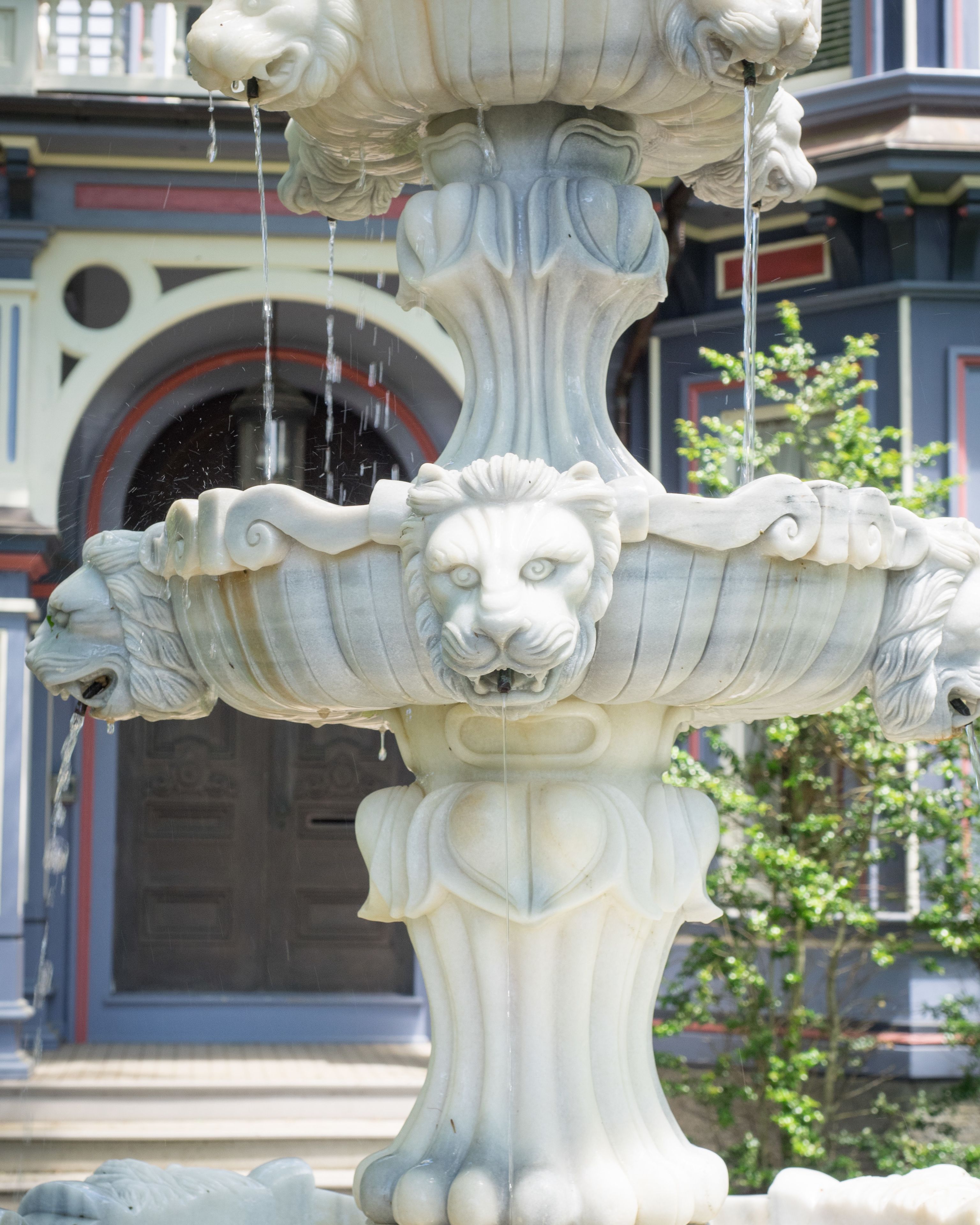 A detailed stone fountain with water flowing from its top, set against a building with a large arched doorway and a tree in the background.