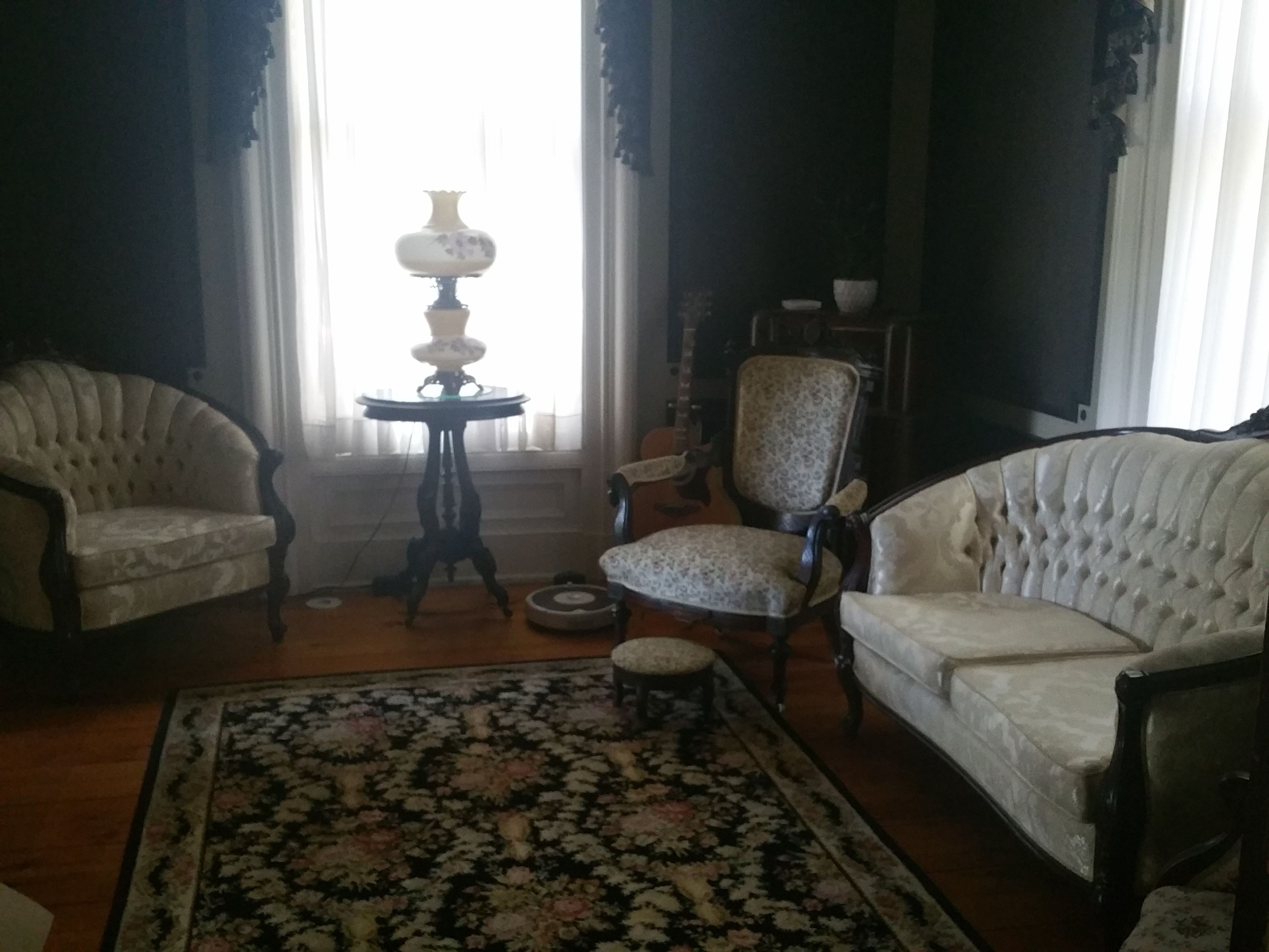 Un salón tradicional con dos sillones tapizados, una mesita redonda con una lámpara, una alfombra estampada y un ventanal con cortinas.