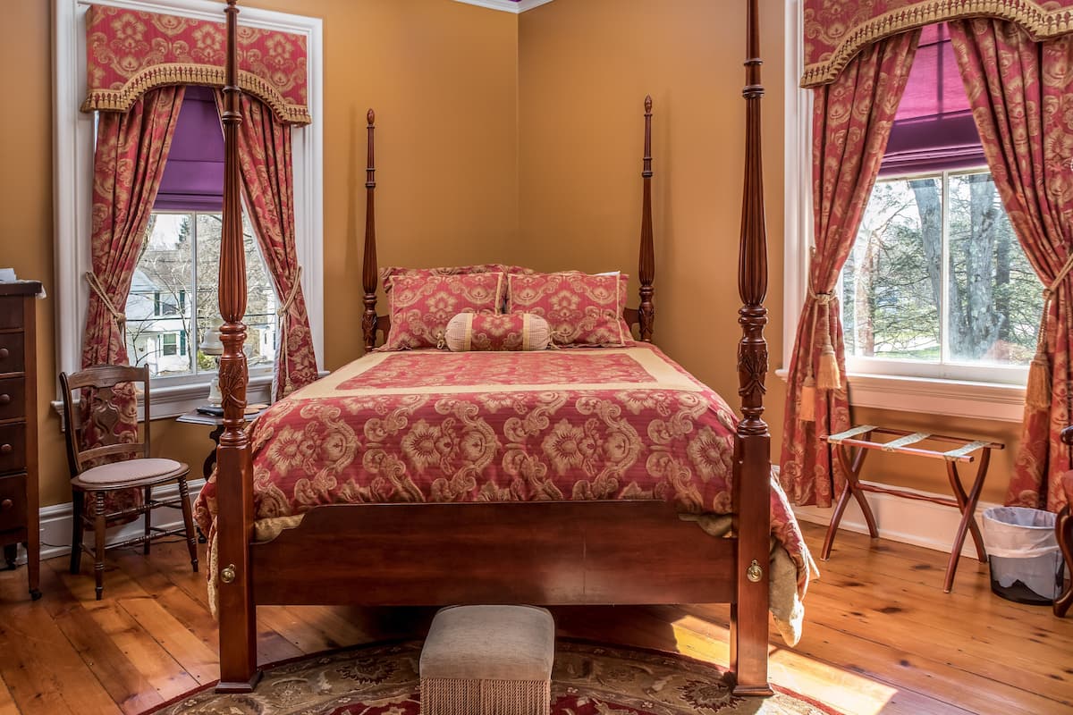 A four-poster bed with a floral bedspread, a wooden dresser, a chair, and a small table.