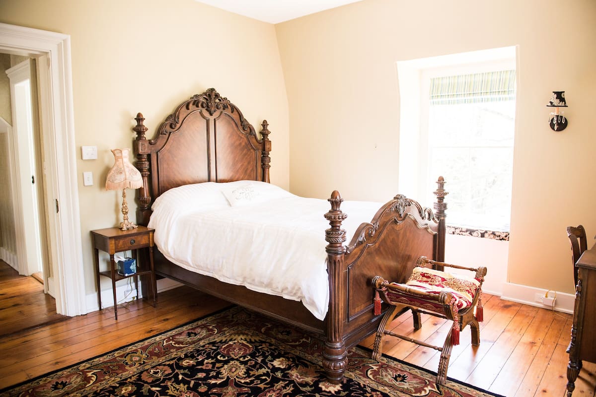A bedroom with a wooden four-poster bed, a nightstand with a lamp, a chair with a red and gold throw, and a large window with a view of trees.