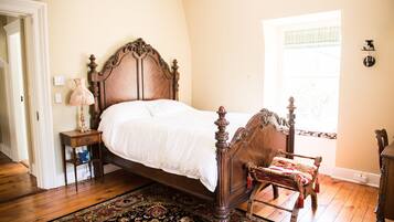 A bedroom with a wooden four-poster bed, a nightstand with a lamp, a chair with a red and gold throw, and a large window with a view of trees.