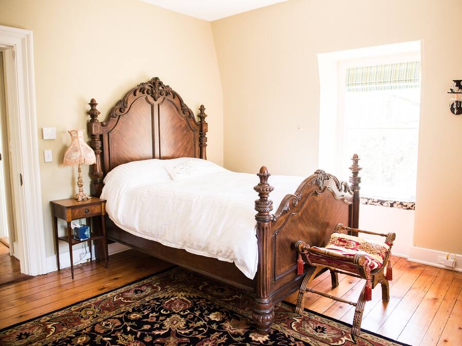 A bedroom with a wooden four-poster bed, a nightstand with a lamp, a chair with a red and gold throw, and a large window with a view of trees.