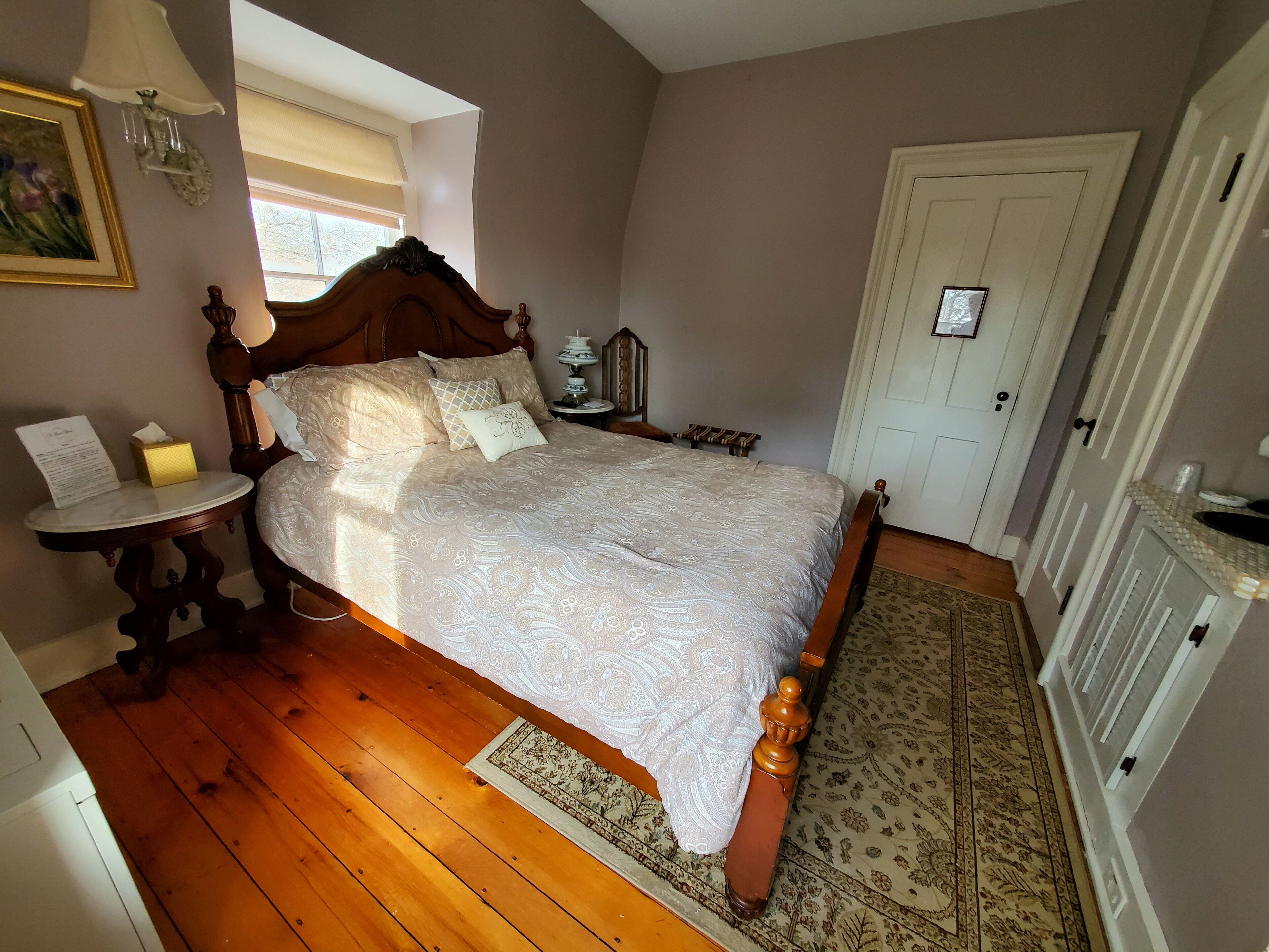 A bedroom with a wooden bed, a bedside table, a nightstand, a lamp, a framed picture on the wall, and a door.