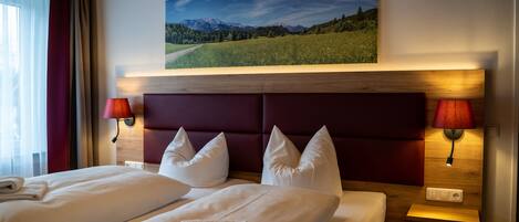 Quarto de hotel com duas camas, cabeceira de madeira, pintura de uma paisagem montanhosa e duas luminárias de cabeceira.