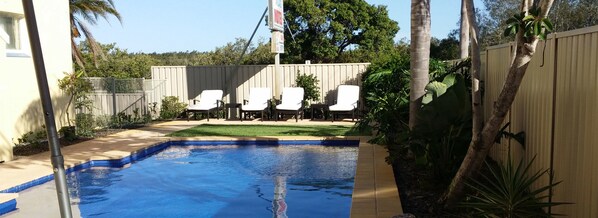 Un patio trasero con piscina, sillas de descanso blancas y un área cercada.