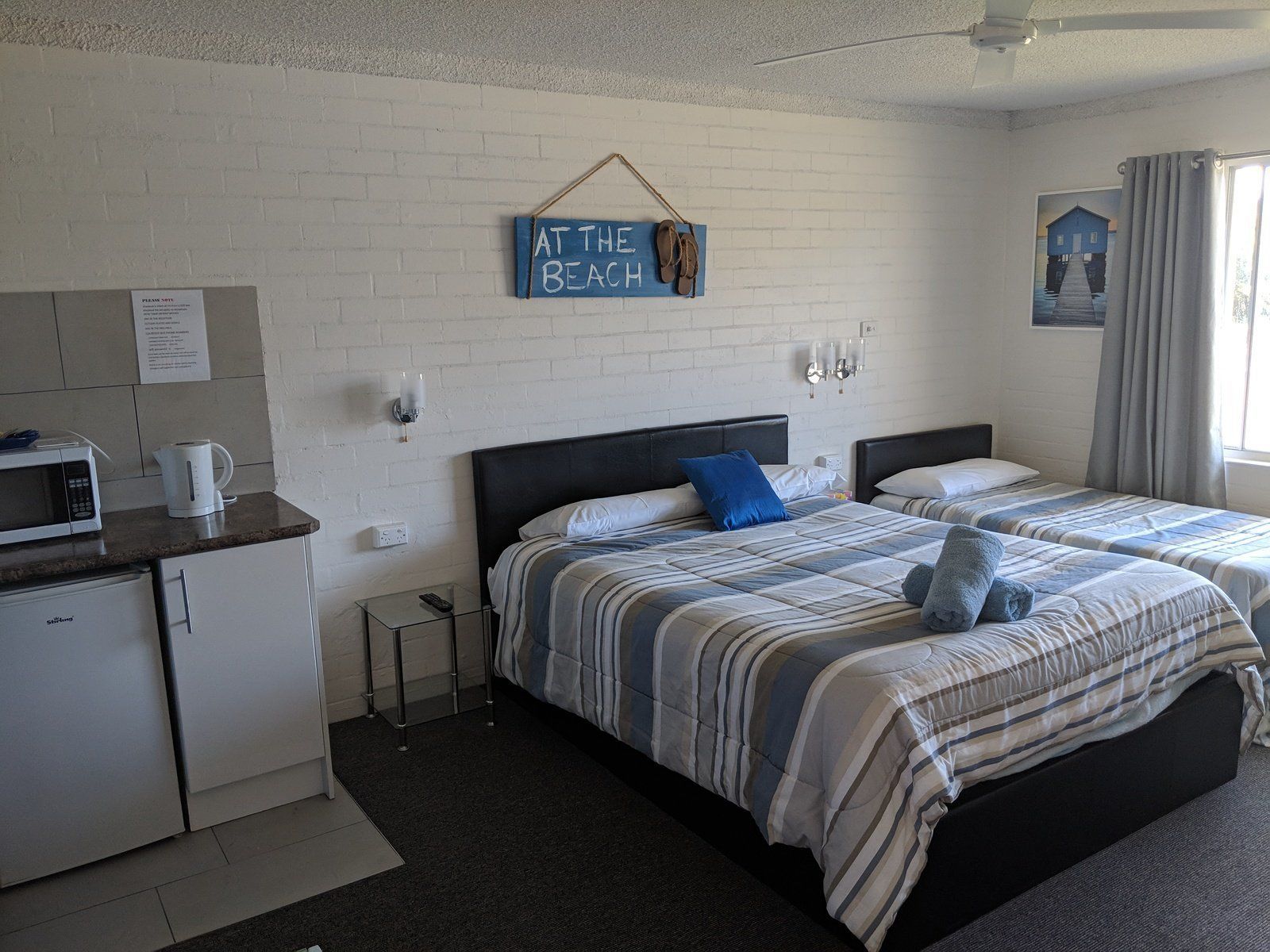 A hotel room with two beds, a kitchenette, a wall-mounted sign reading "AT THE BEACH", and a window with curtains.
