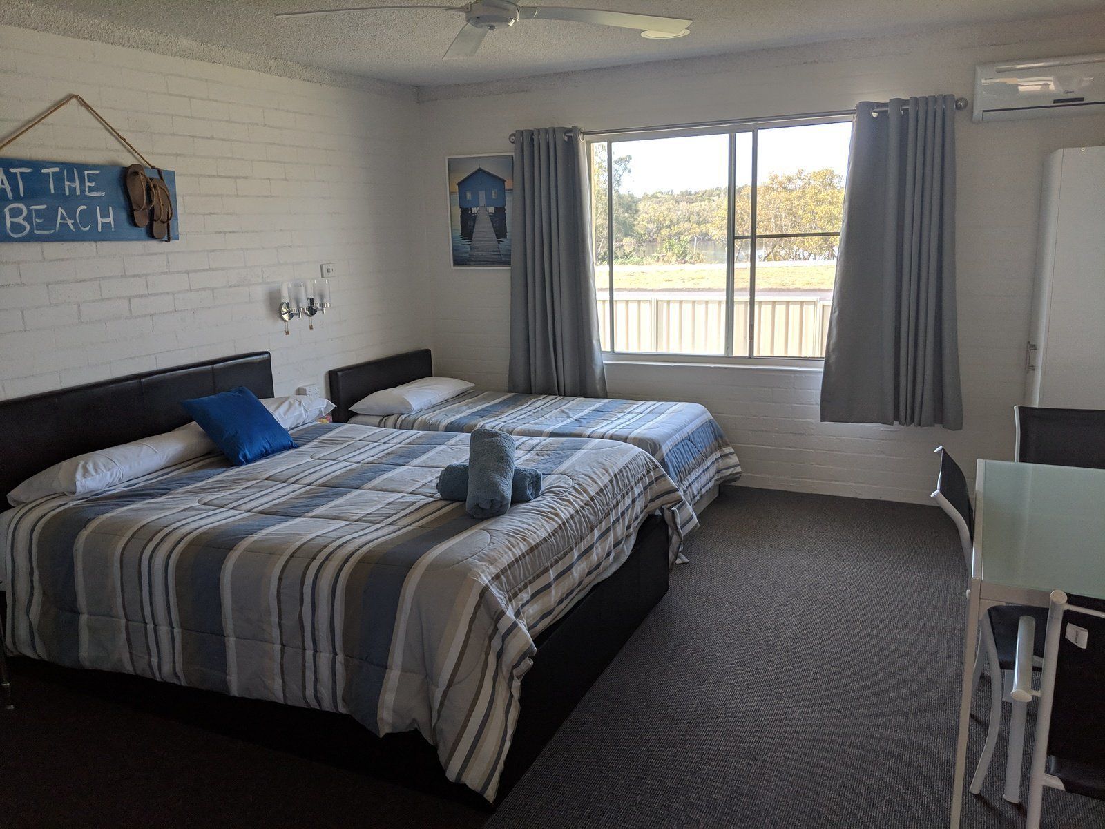 A hotel room with two beds, a desk, a chair, a window with curtains, a wall-mounted picture, and a sign reading "AT THE BEACH".