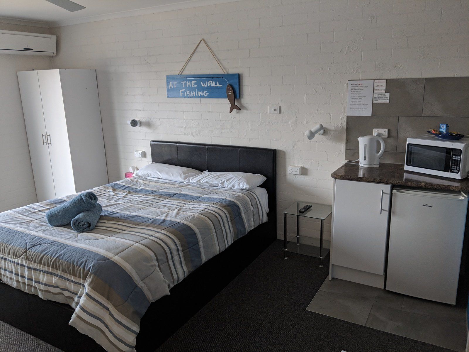 A hotel room with a bed, a small kitchenette, a white wardrobe, and a wall-mounted sign that reads "AT THE WALL FISHING".