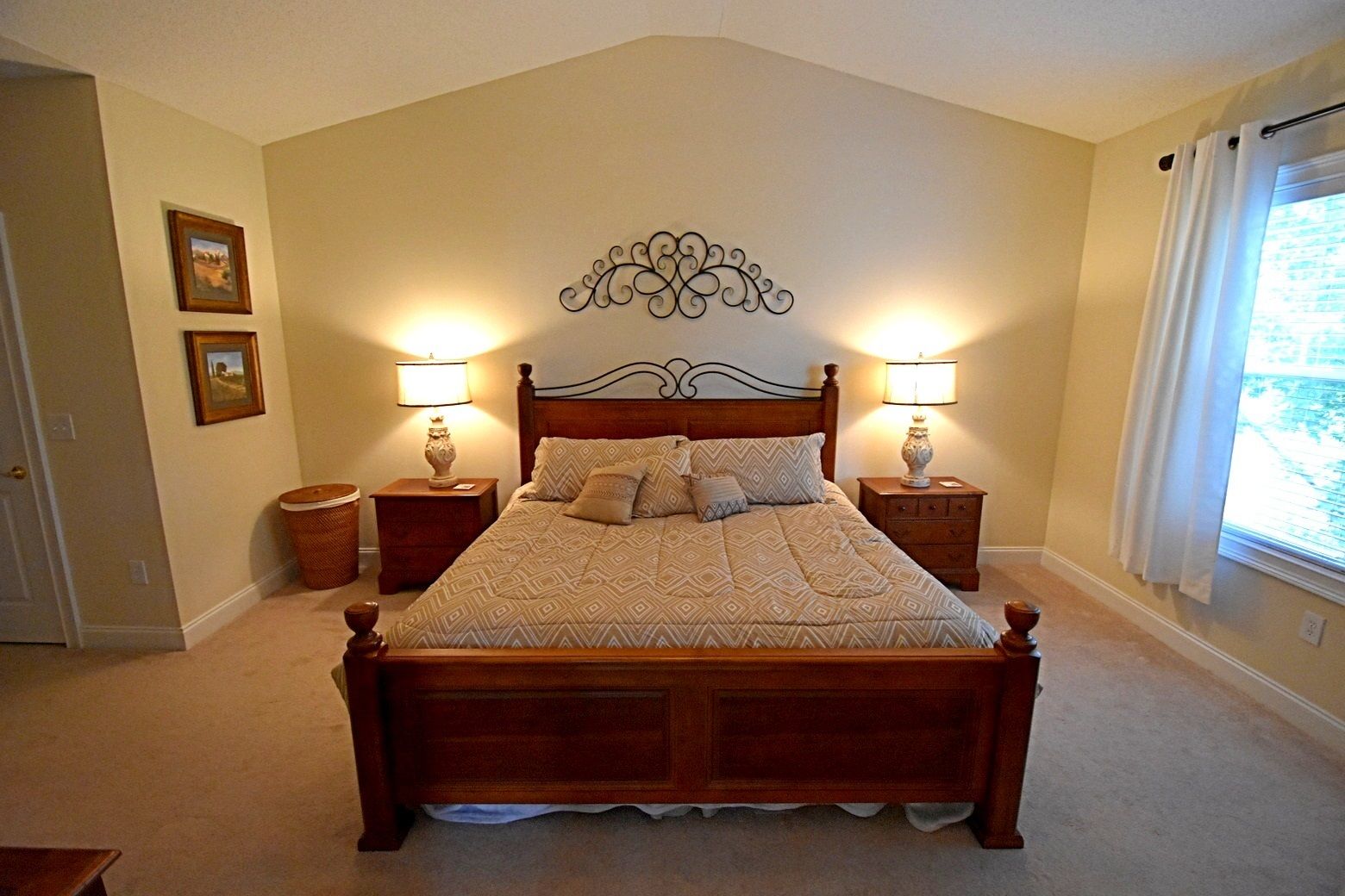Un dormitorio con una cama de madera, dos mesitas de noche con lámparas, un adorno en la pared y una ventana con cortinas.