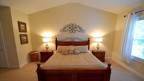 A bedroom with a wooden bed, two bedside tables with lamps, a decorative wall piece, and a window with curtains.
