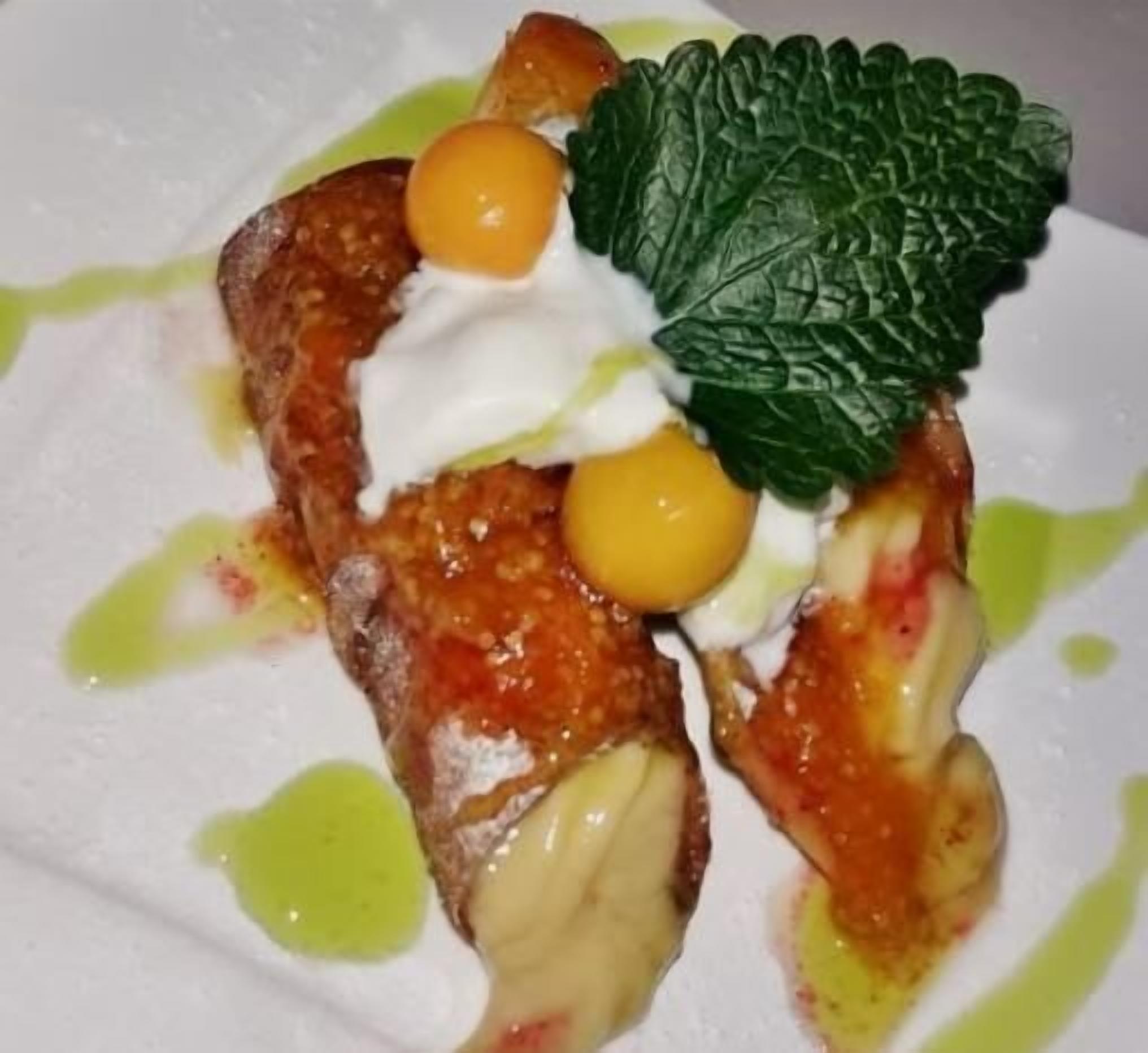 Un postre con cobertura cremosa, dos esferas amarillas y una hoja de menta sobre un plato blanco con salsa verde.