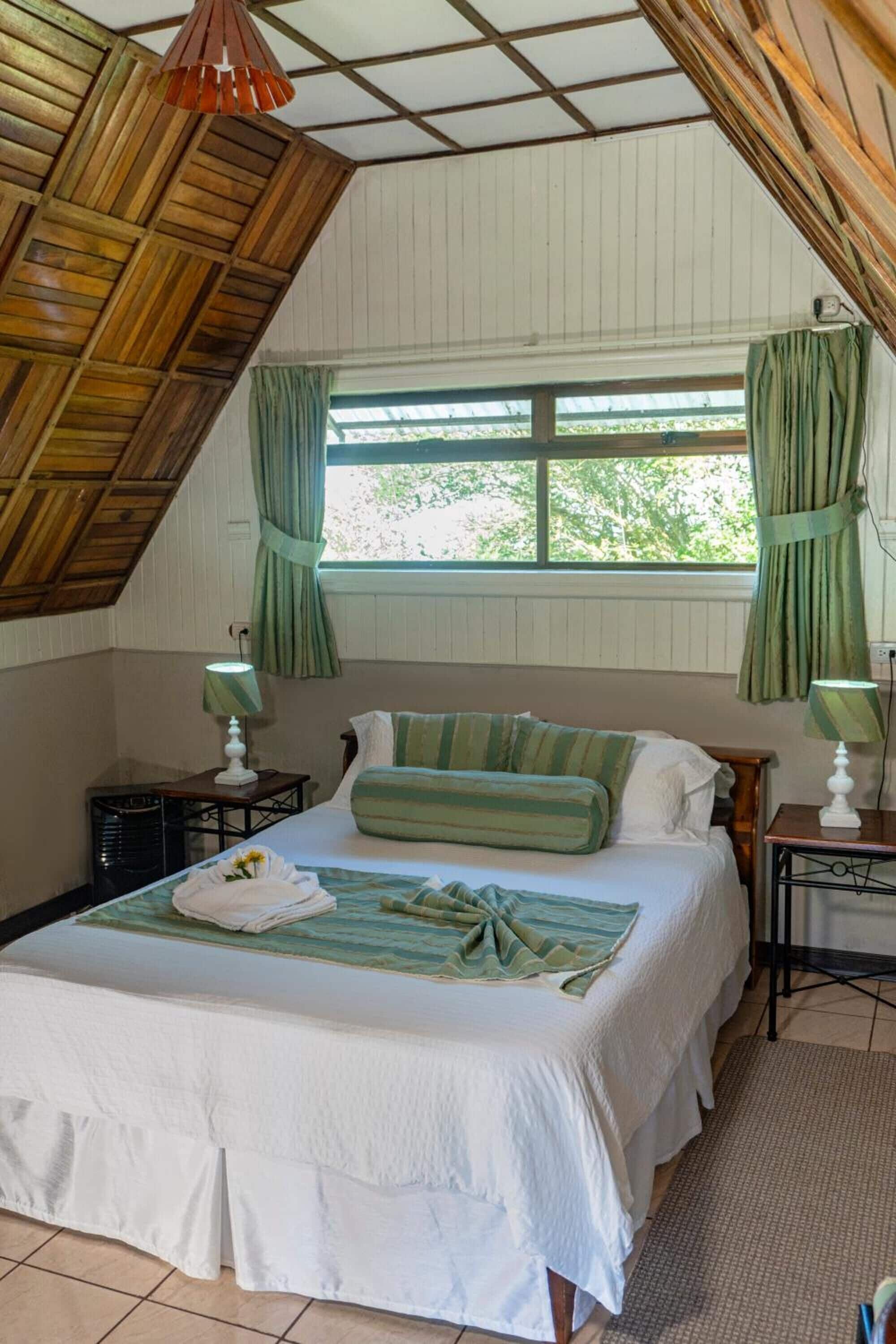Un dormitorio con cama, dos mesitas de noche, una ventana con cortinas y un techo inclinado.