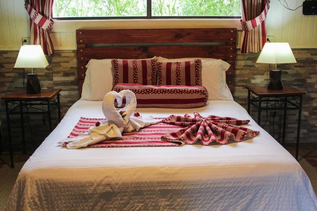 A neatly made bed with a red and white patterned blanket, a towel, and a pillow with a similar pattern. There are two bedside tables with lamps, and a window with curtains.