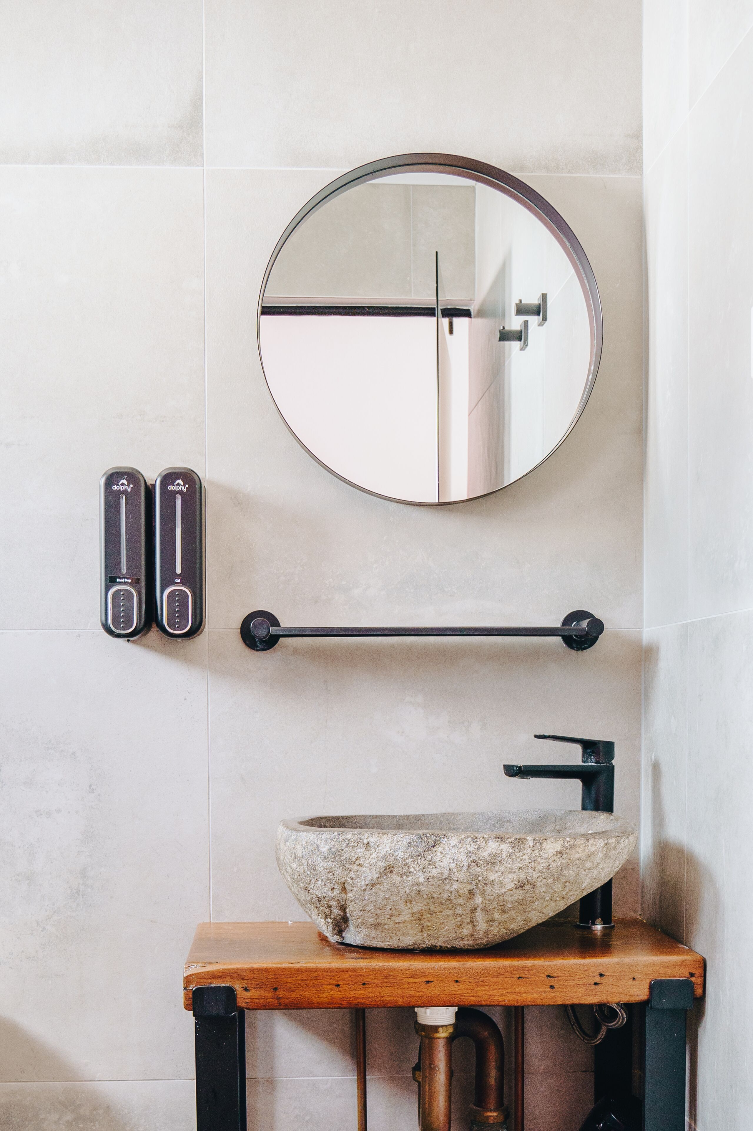 A bathroom with a round mirror, a stone sink, and two soap dispensers.