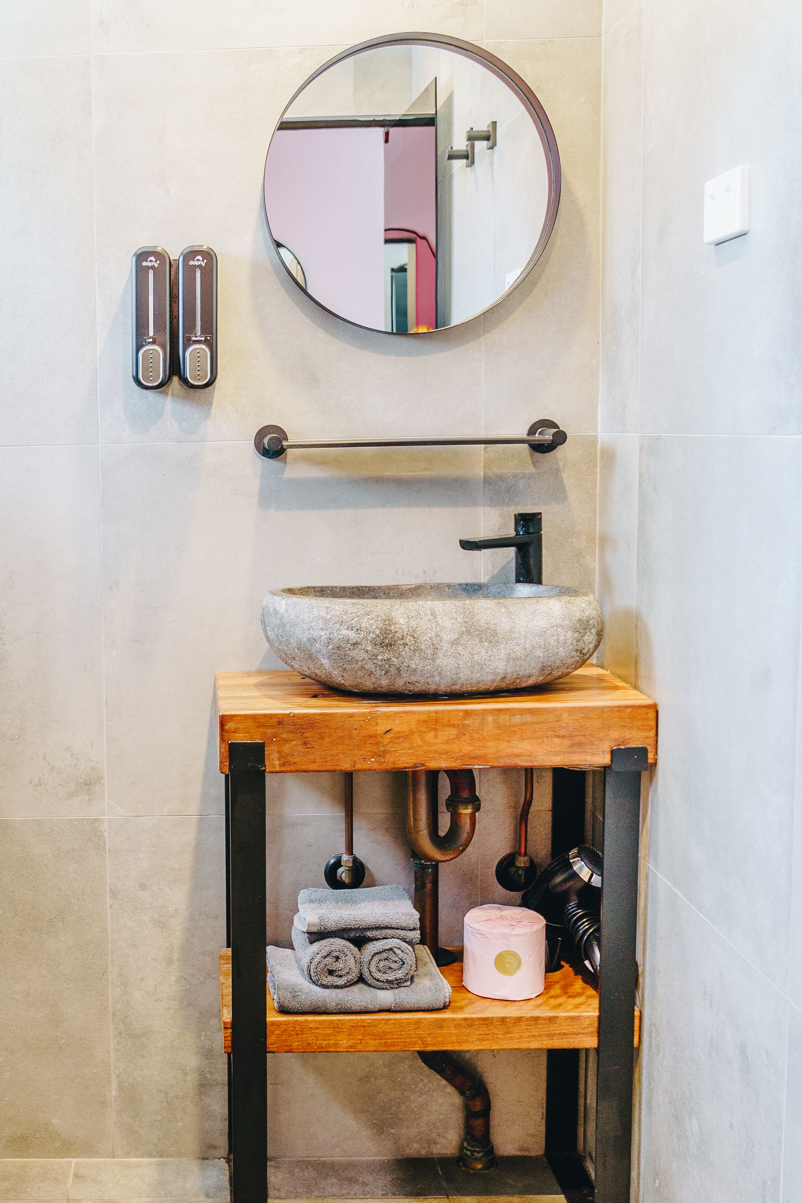 A bathroom with a round mirror, a stone sink, a towel rack, and a shelf with towels and a candle.