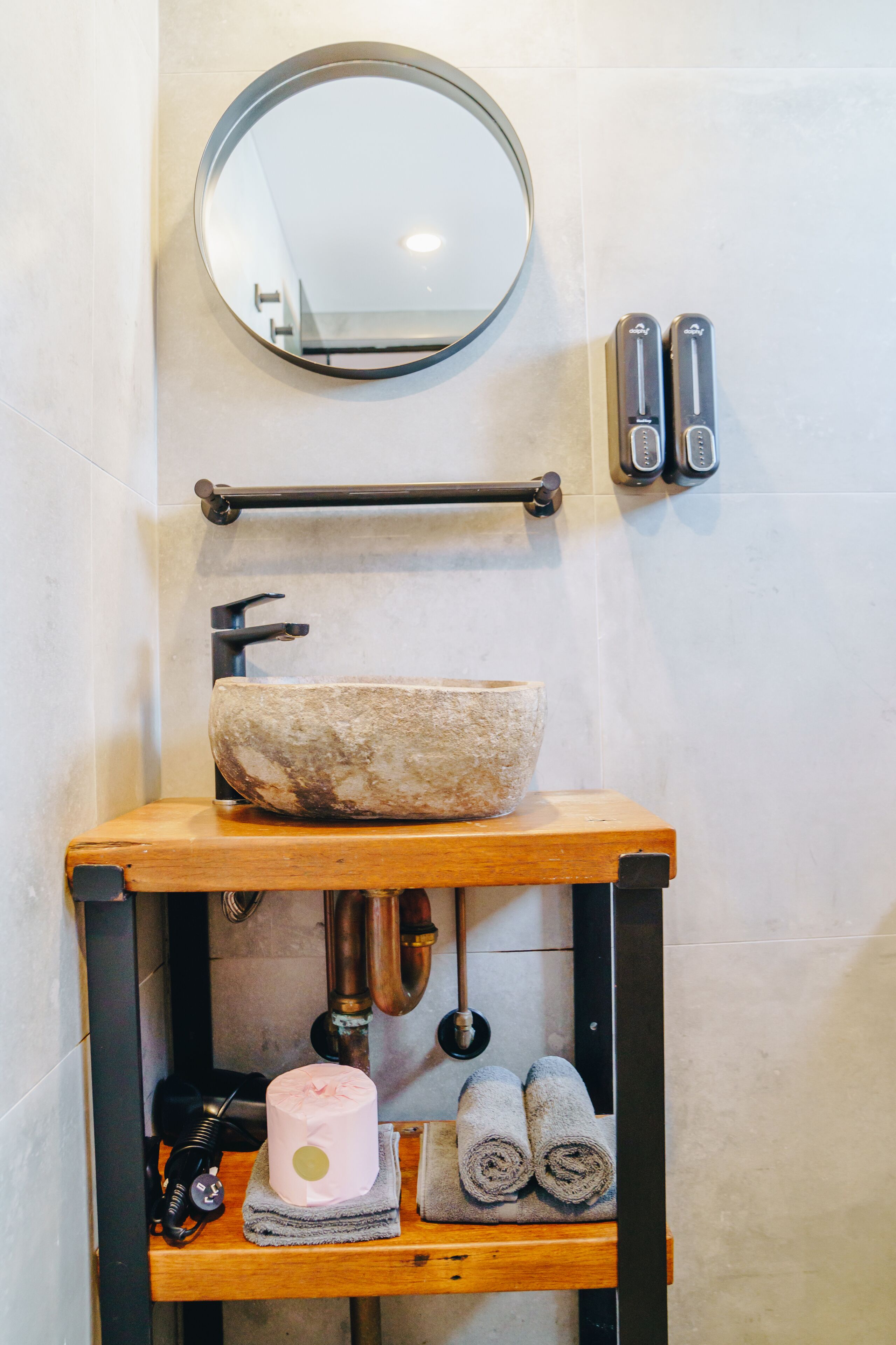 A bathroom with a stone sink, a round mirror, towel racks, and a wooden shelf with towels and a cake.