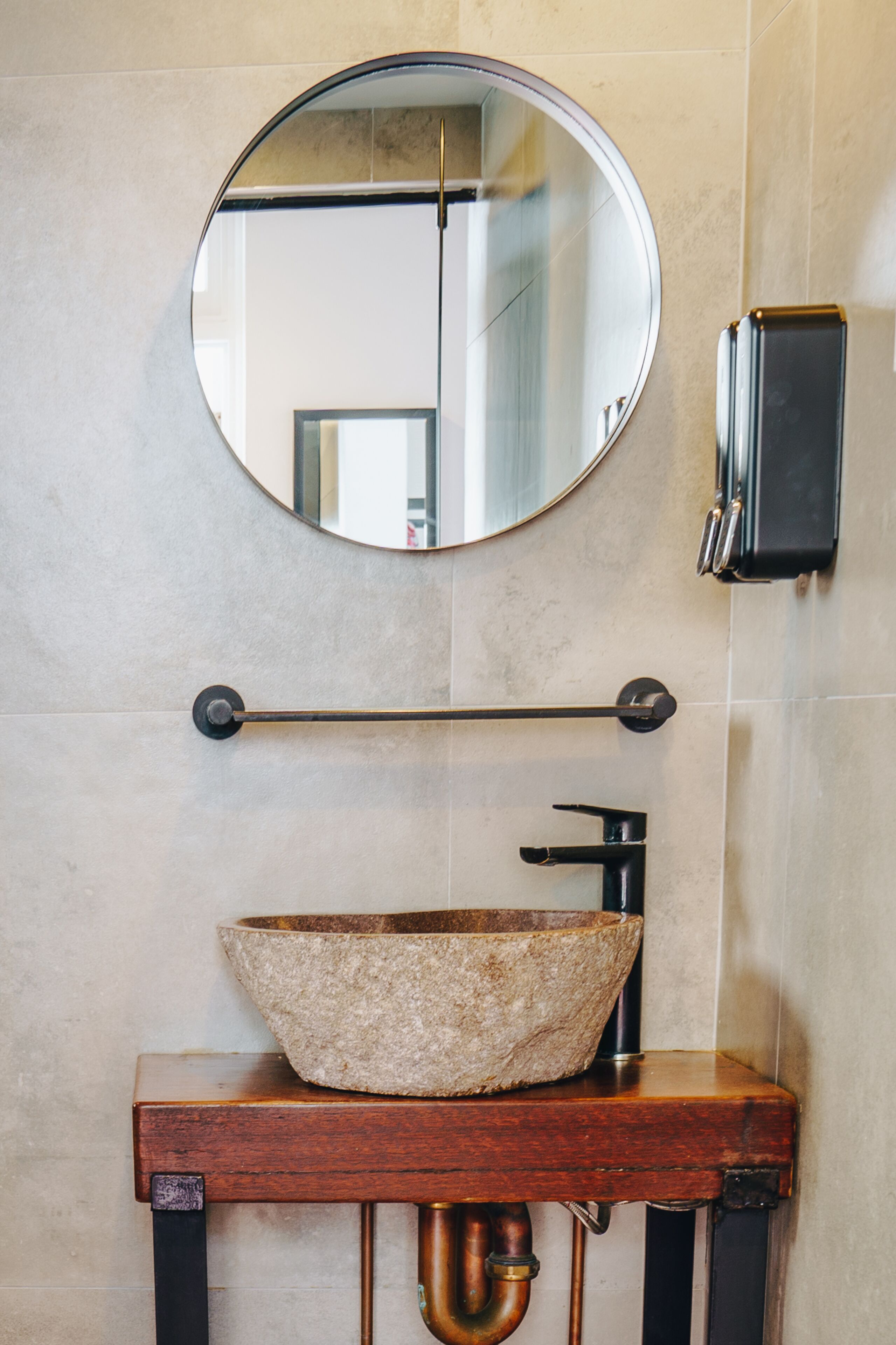 Een badkamer met een ronde spiegel, een houten wastafelmeubel met een stenen wasbak, een handdoekrek en een zeepdispenser.