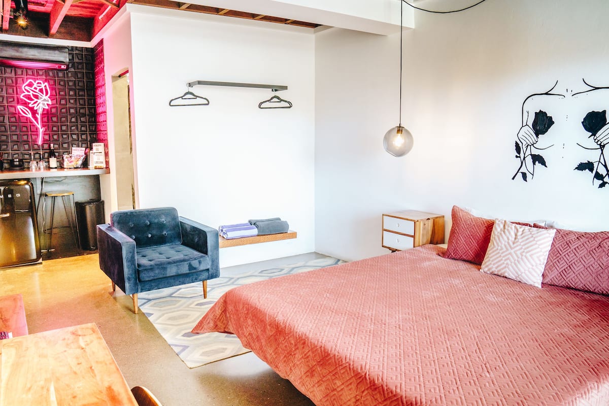Een moderne slaapkamer met een groot bed, een blauwe fauteuil, een klein bureau en een neon bord met een bloem.